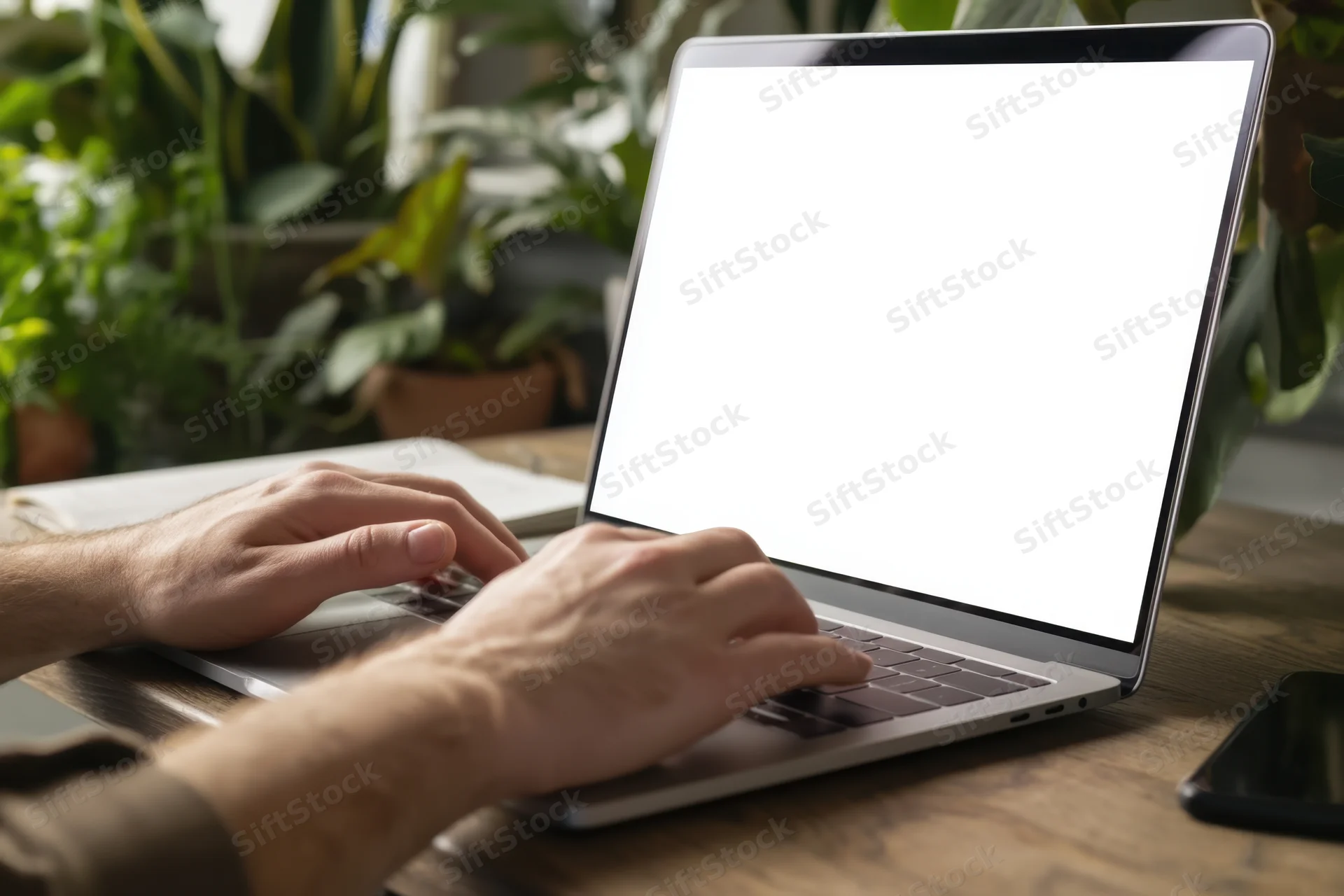 Man typing on a modern laptop with blank screen on wooden desk surrounded by green plants