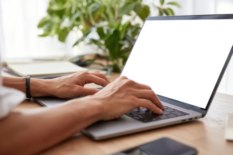 Woman typing on a laptop with blank screen at a wooden desk in a bright indoor workspace