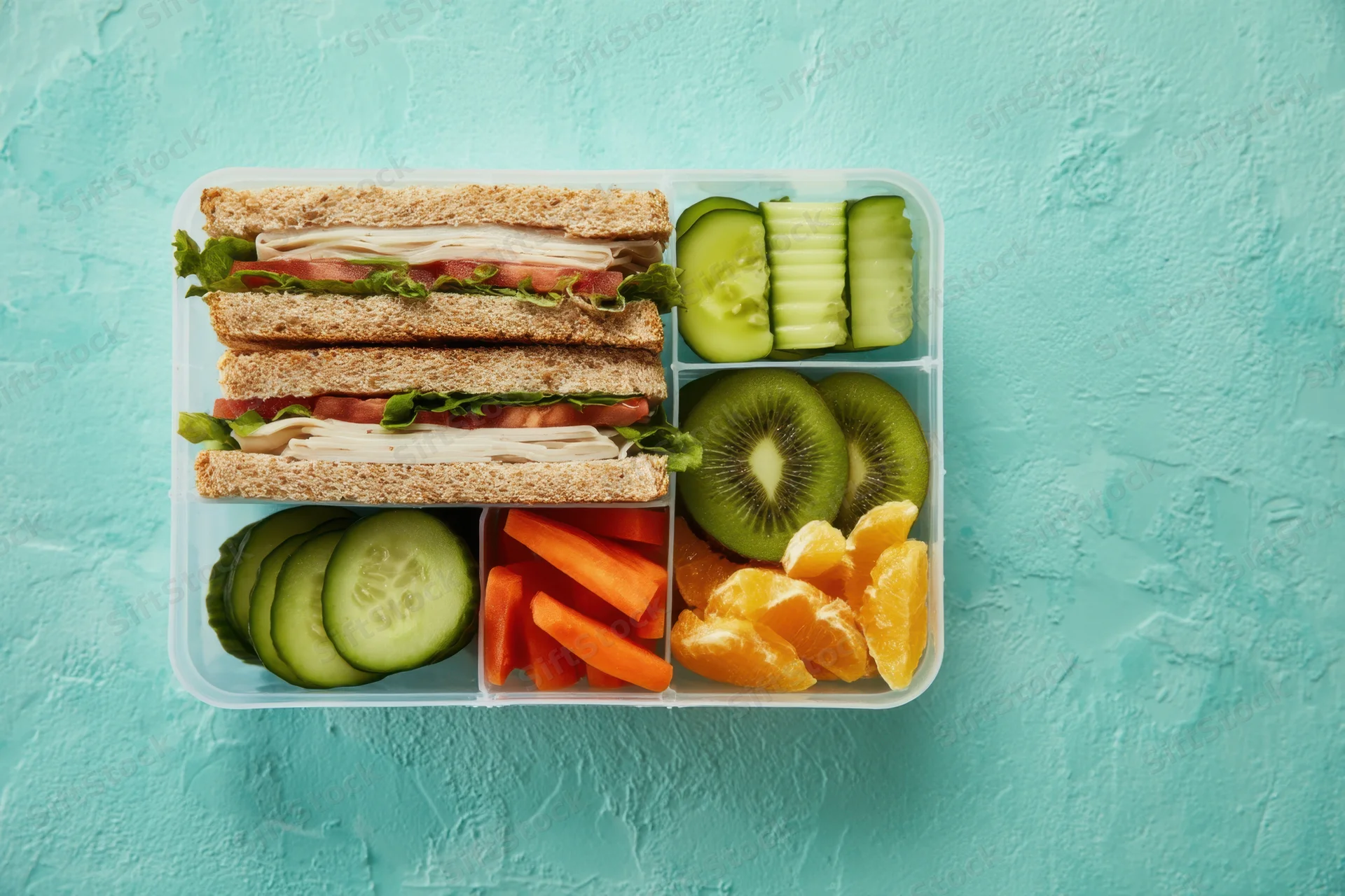 Compartmentalized lunchbox with sandwiches, sliced fruits and vegetables arranged on a teal background