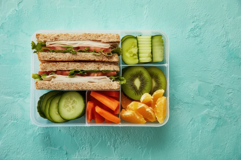 Compartmentalized lunchbox with sandwiches, sliced fruits and vegetables arranged on a teal background