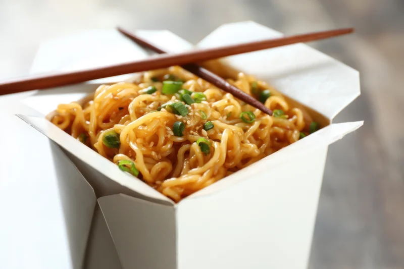 Golden noodles with green onions and sesame in a white takeout box with chopsticks resting on top
