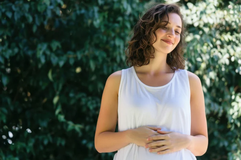 Woman in white sleeveless top standing outdoors with hands on stomach smiling