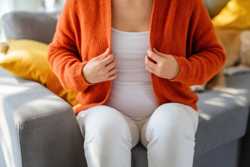 Pregnant woman seated on sofa wearing orange cardigan over white top and white pants with yellow cushions nearby