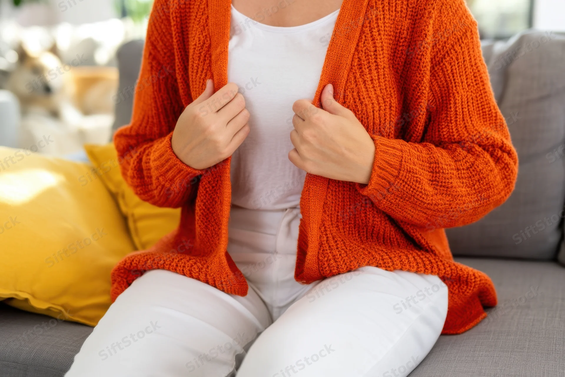 Woman in orange cardigan over white top seated on gray sofa holding garment edges