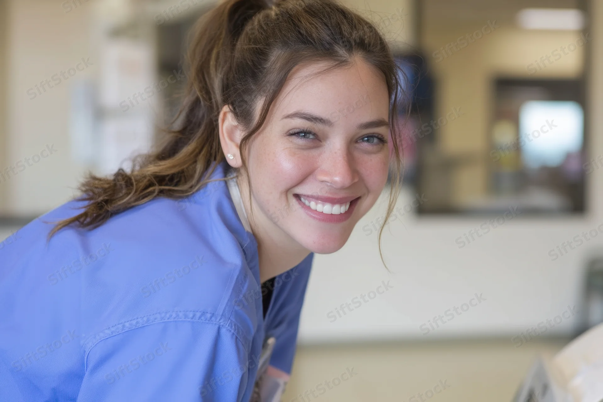 Smiling clinician in blue scrubs with ponytail leaning in a medical facility
