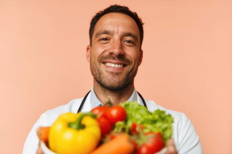 Smiling male doctor in lab coat holding bowl of fresh vegetables against peach background