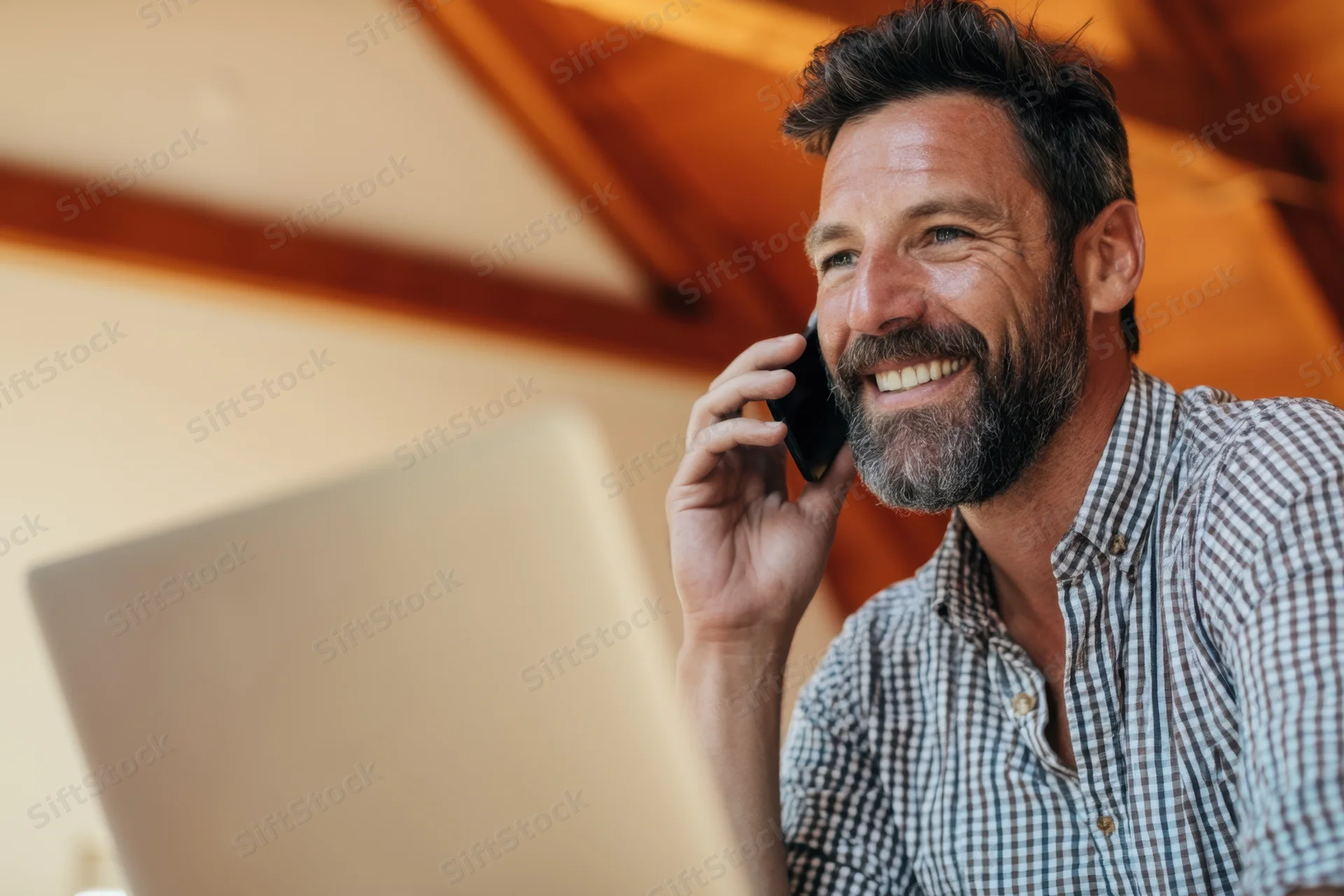 Smiling man in checkered shirt talking on phone while working at laptop in home office
