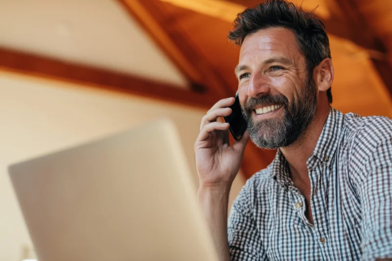 Smiling man in checkered shirt talking on phone while working at laptop in home office