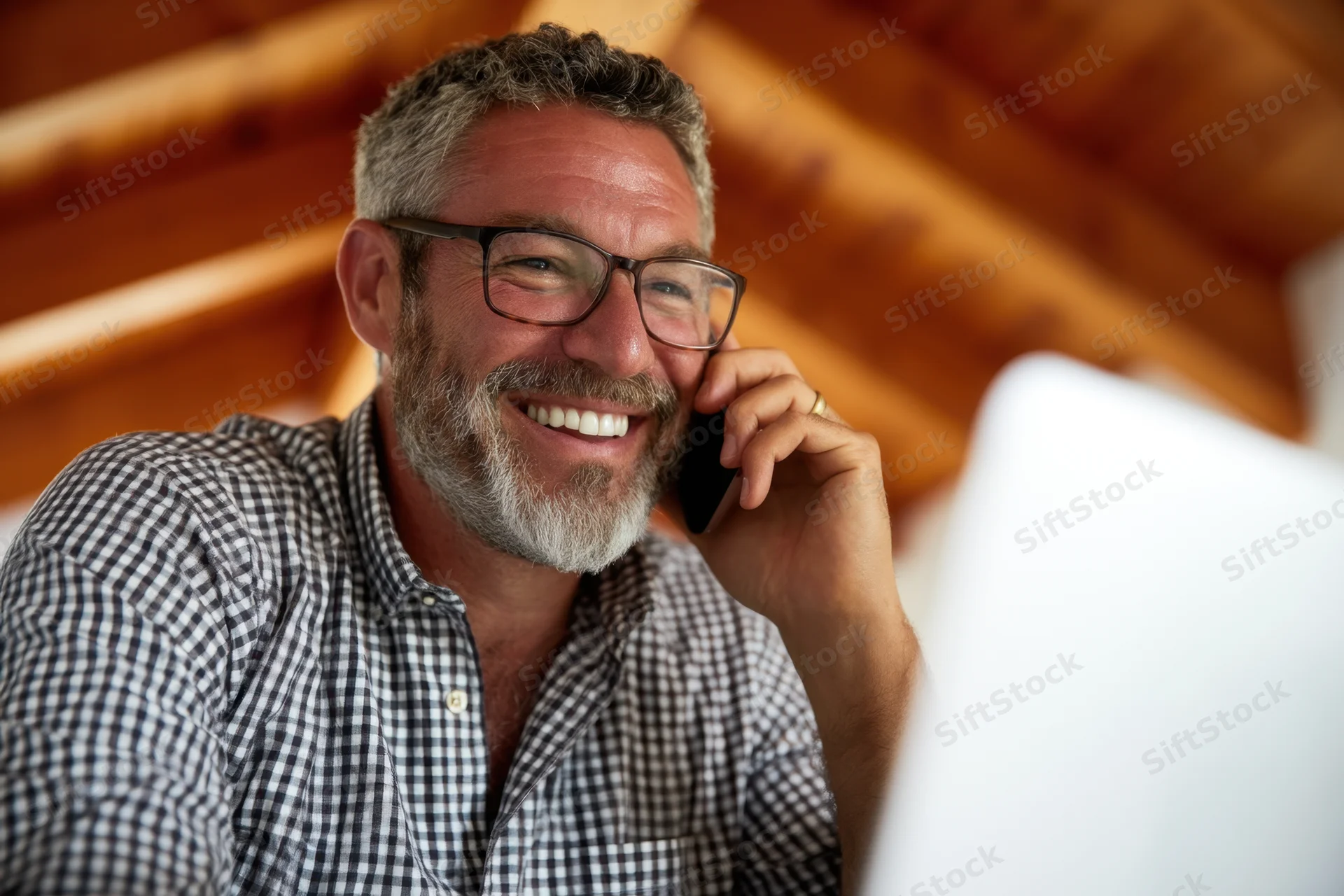Smiling man in checkered shirt talking on phone while working at laptop in home office