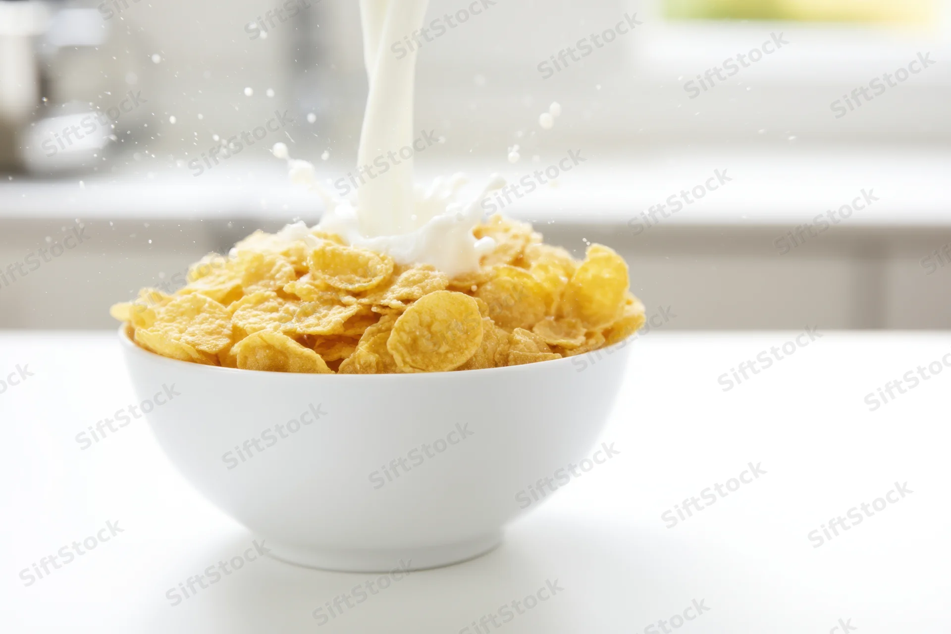 Milk being poured into a white ceramic bowl filled with golden cornflakes on a kitchen table