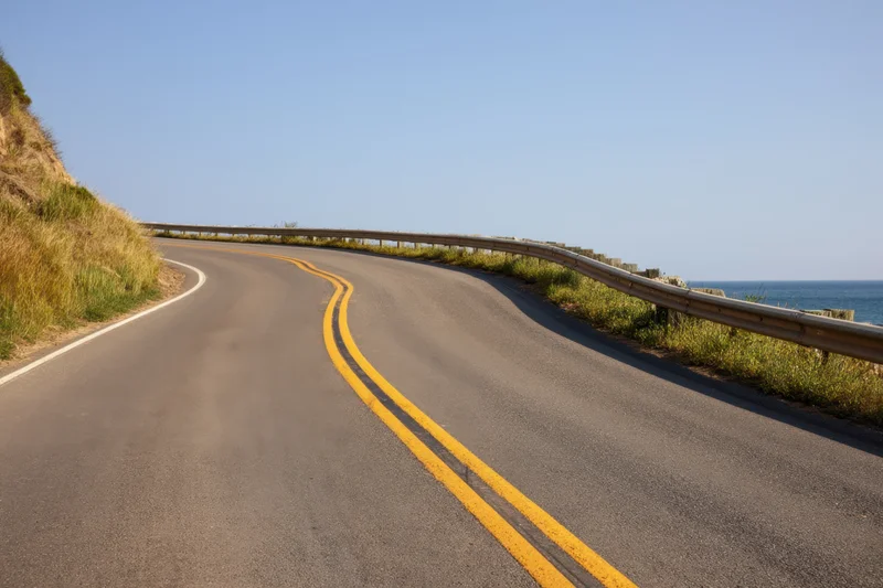 Winding asphalt road curving along a coastal cliff with ocean view and guardrail