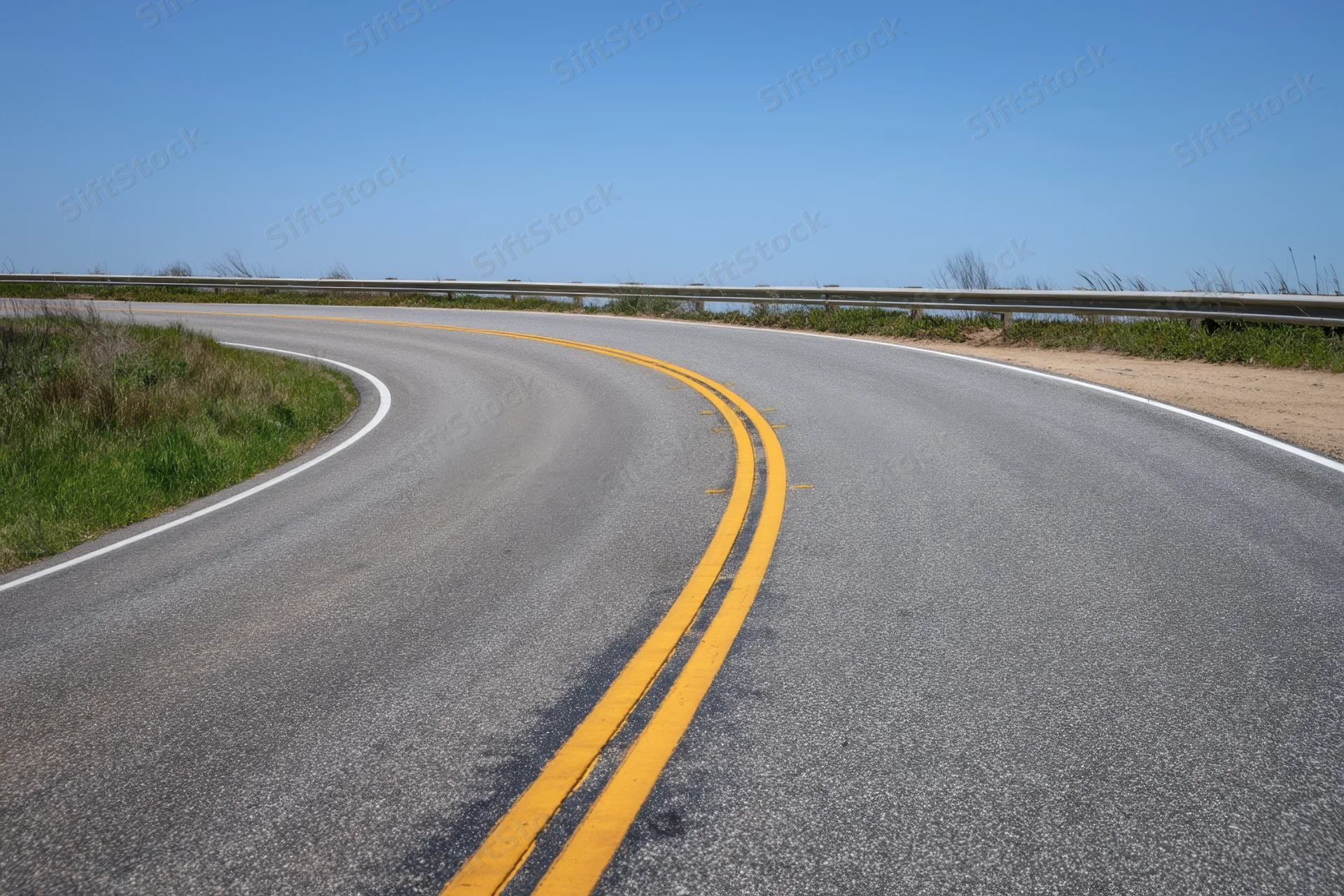 Winding asphalt road with double yellow lines curving along a coastal guardrail under clear blue sky