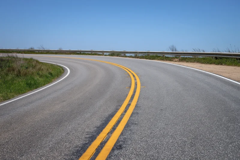 Winding asphalt road with double yellow lines curving along a coastal guardrail under clear blue sky