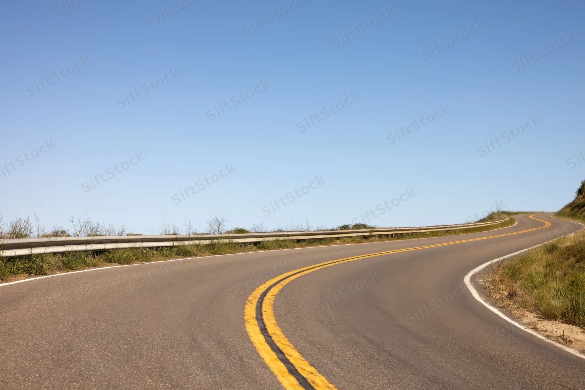 Winding asphalt road curving through rural landscape with yellow painted lines and guardrail under clear blue sky