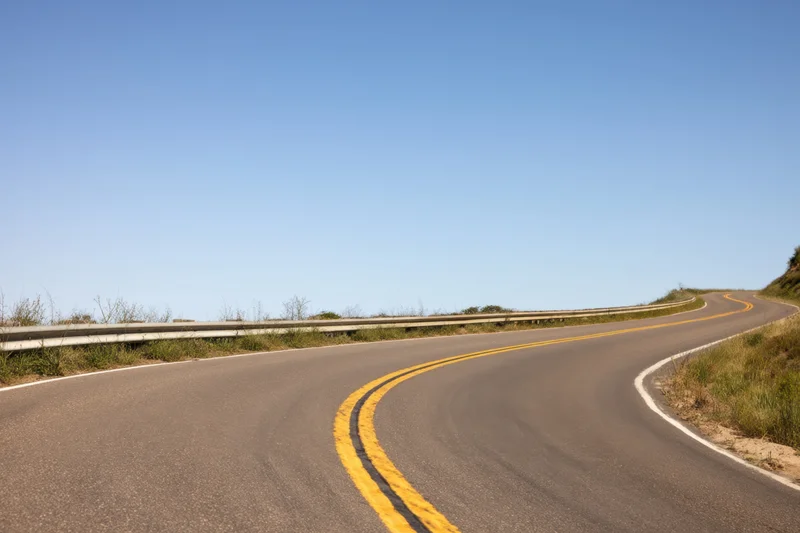 Winding asphalt road curving through rural landscape with yellow painted lines and guardrail under clear blue sky
