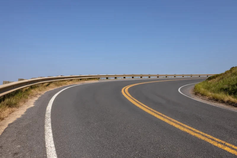 Winding asphalt road curving through rural landscape with guardrail and painted lane markings under clear blue sky
