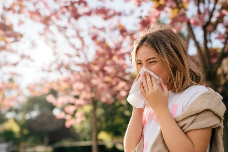 Young woman sneezing into tissue in a cherry blossom park during spring
