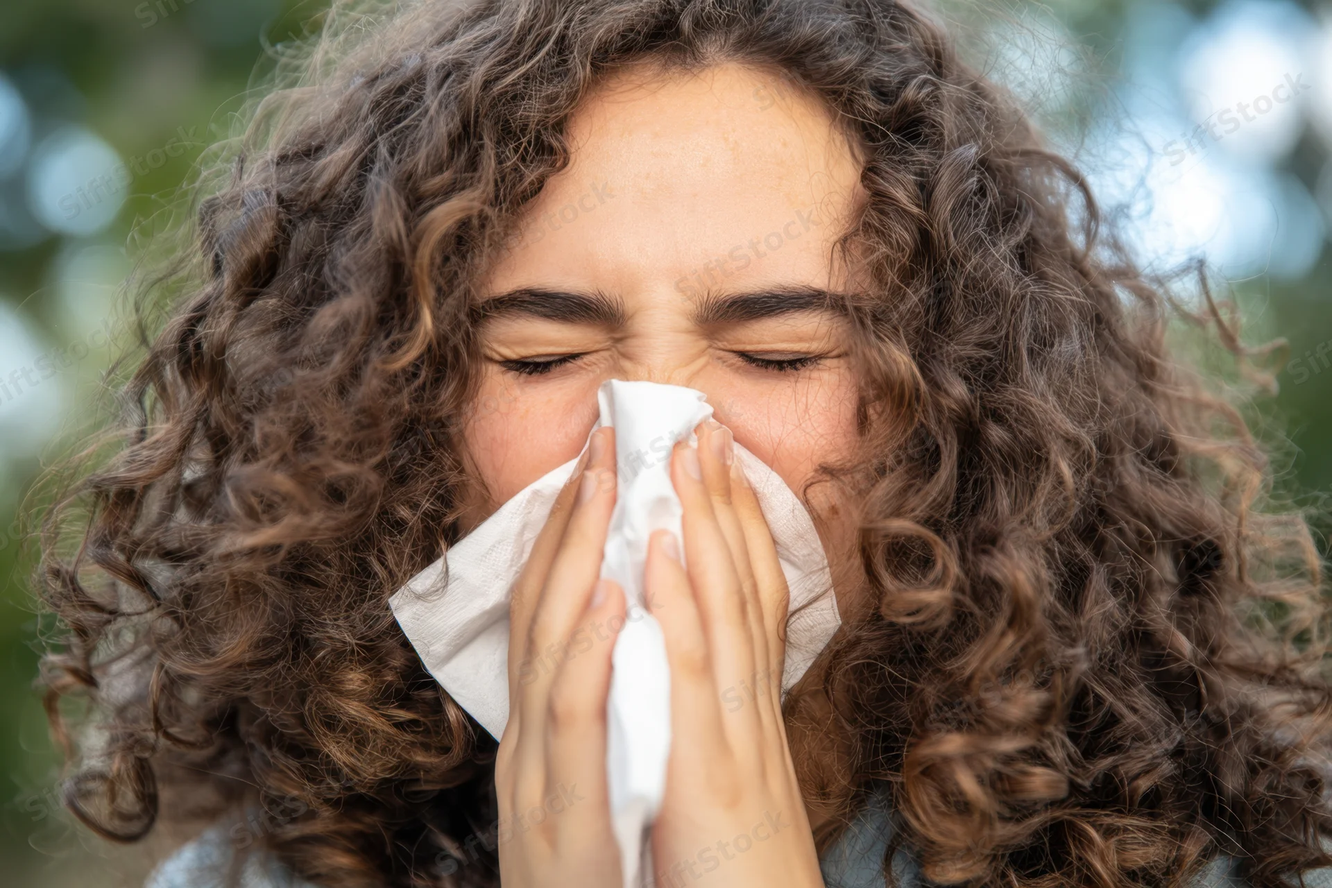 Young woman with curly hair sneezing into tissue outdoors