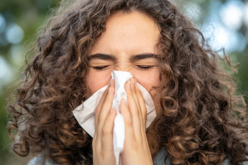 Young woman with curly hair sneezing into tissue outdoors