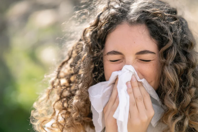 Young woman with curly hair sneezing into a tissue outdoors in a green setting
