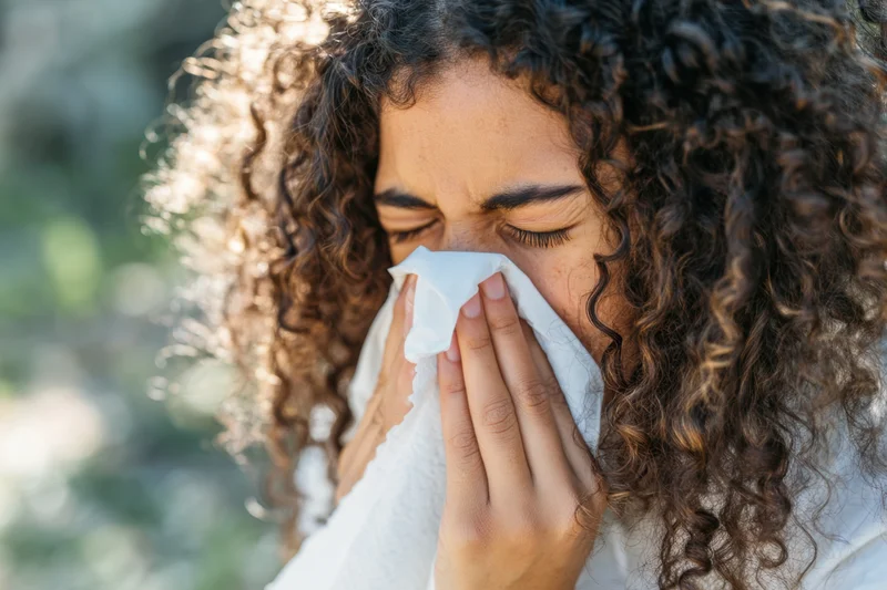 Young woman with curly hair sneezing into tissue outdoors in natural light