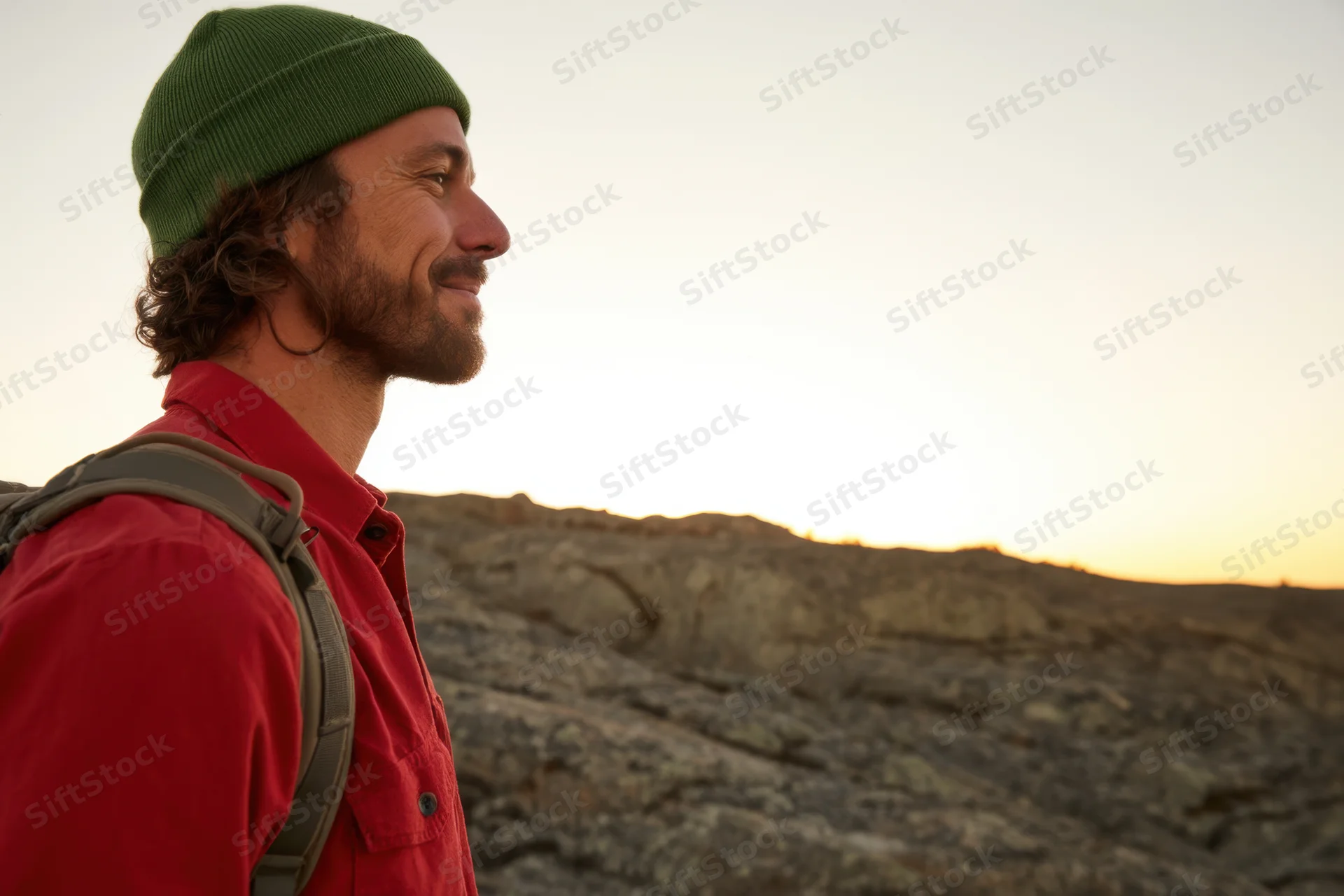 Young man smiling in profile on rocky terrain at sunset wearing red shirt and green beanie with backpack