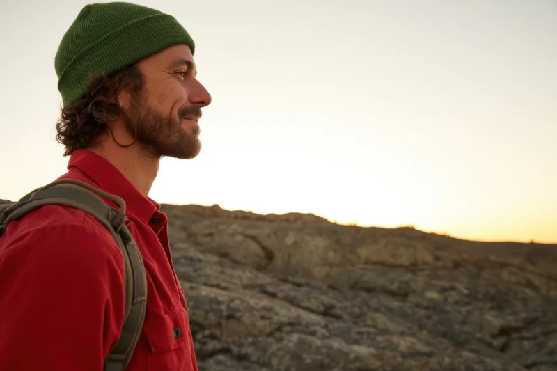 Young man smiling in profile on rocky terrain at sunset wearing red shirt and green beanie with backpack