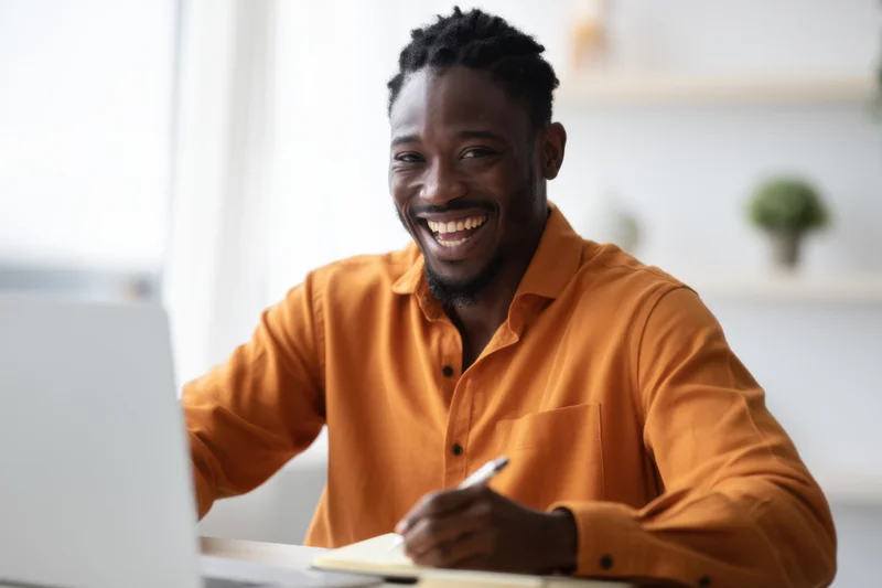 Young black man laughing while writing in notebook at desk with laptop open in front