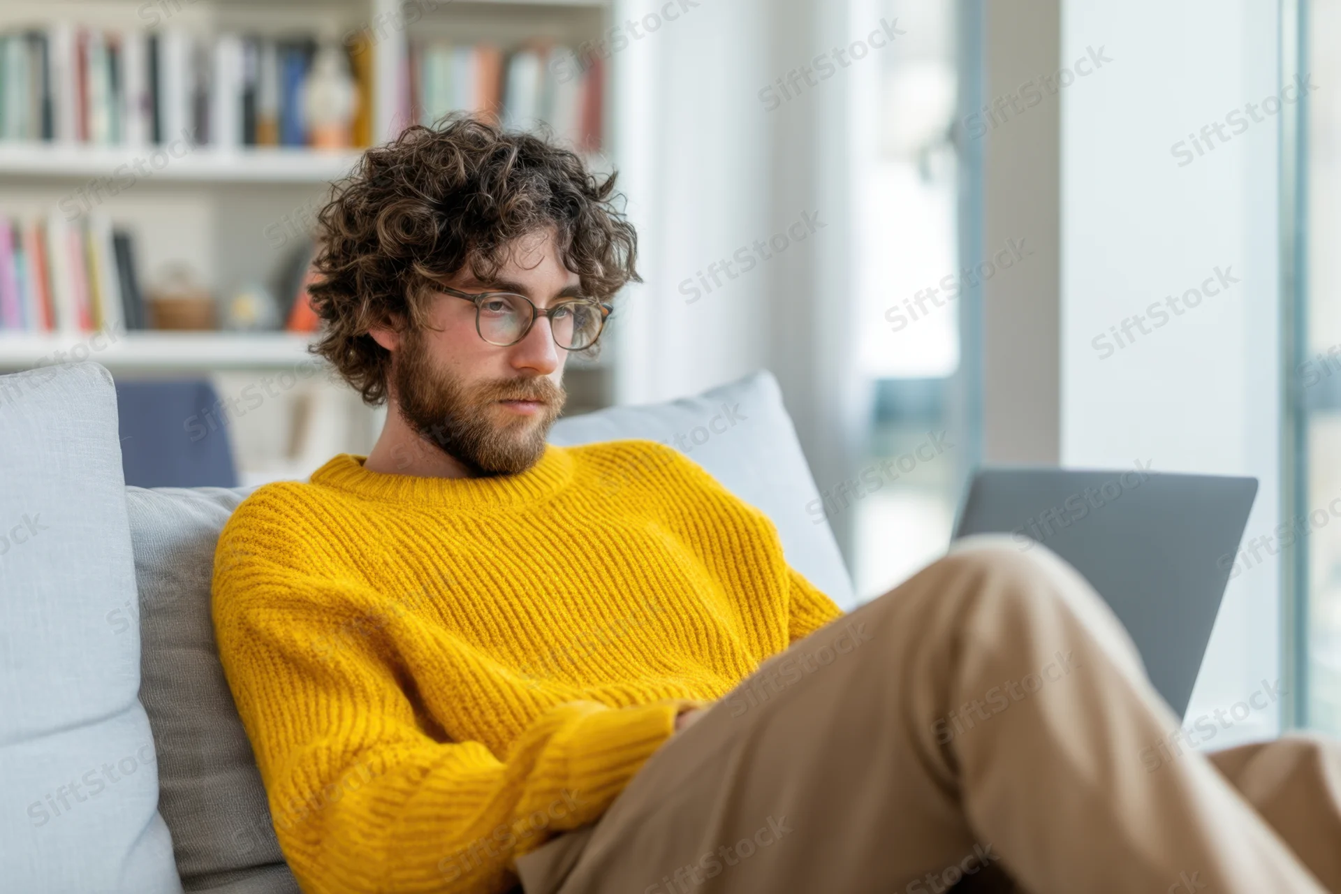 Young man with curly hair and glasses focused on laptop while sitting on couch in home office setting