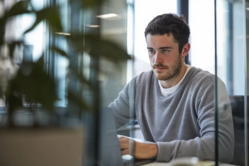 Young man concentrating on laptop at office desk behind glass partition