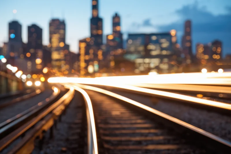 Train tracks with light trails leading toward a blurred city skyline at dusk
