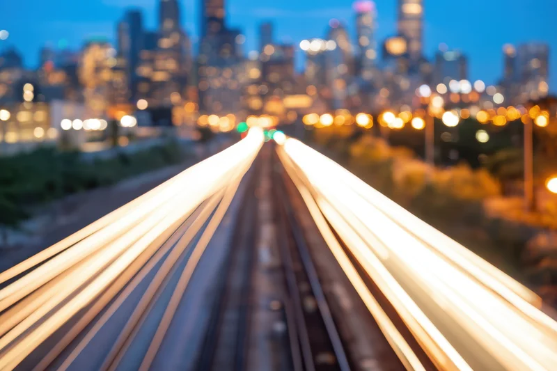 Train tracks with light streaks leading toward a blurred city skyline at dusk