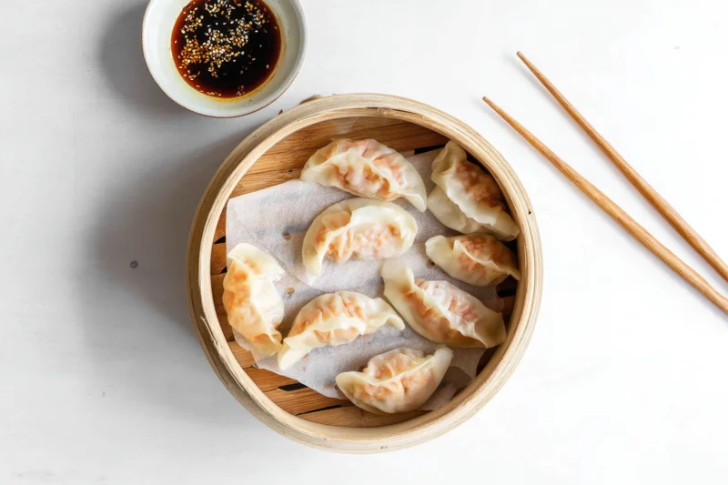 Bamboo steamer filled with dumplings on white surface with chopsticks and dipping sauce nearby