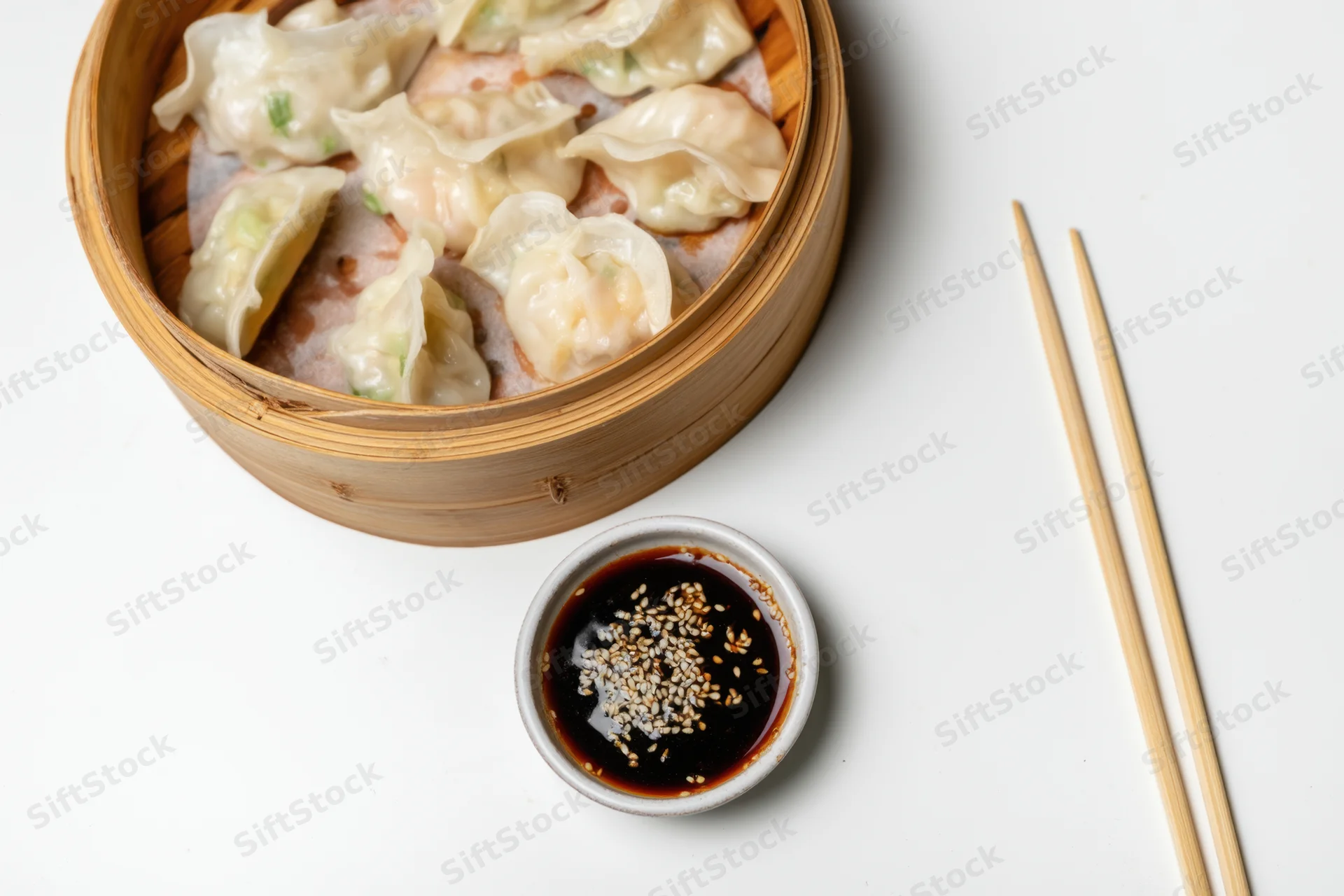 Bamboo steamer filled with dumplings and soy sauce on white surface with chopsticks