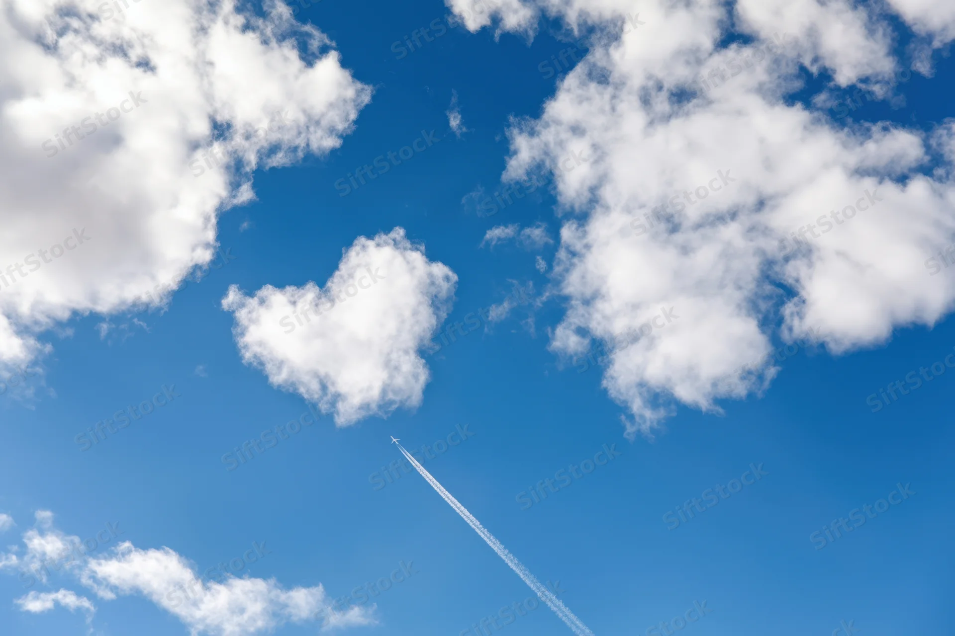 Airplane flying across bright blue sky leaving contrail among fluffy white clouds