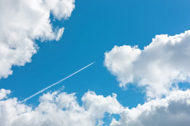 Airplane flying across a bright blue sky leaving a white contrail between fluffy white clouds