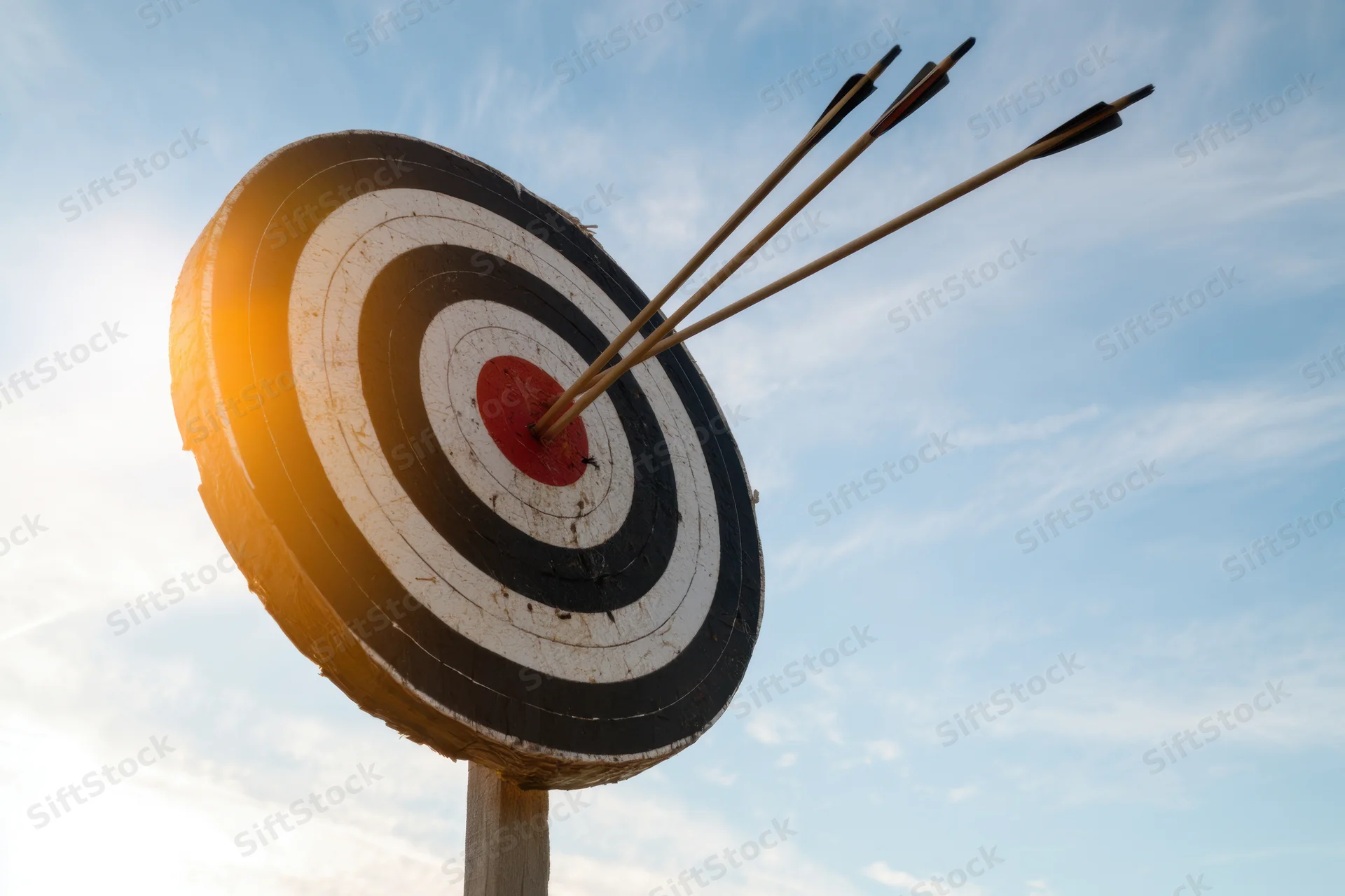 Bullseye target with three arrows embedded in the center against a blue sky with sunlight