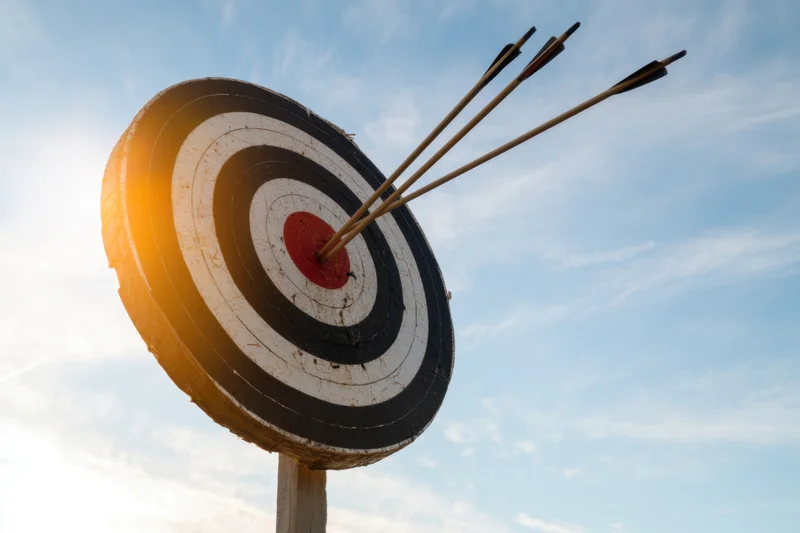 Bullseye target with three arrows embedded in the center against a blue sky with sunlight