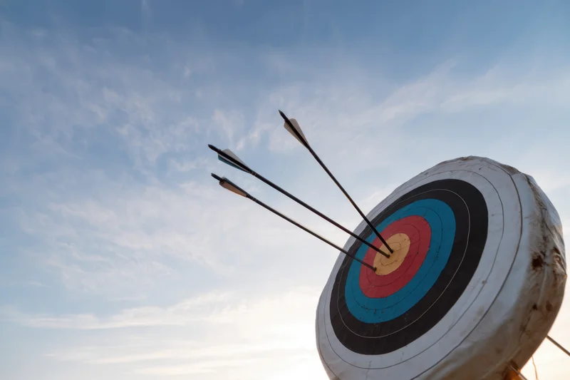 Three arrows embedded in the center of a bullseye target against a blue sky with scattered clouds