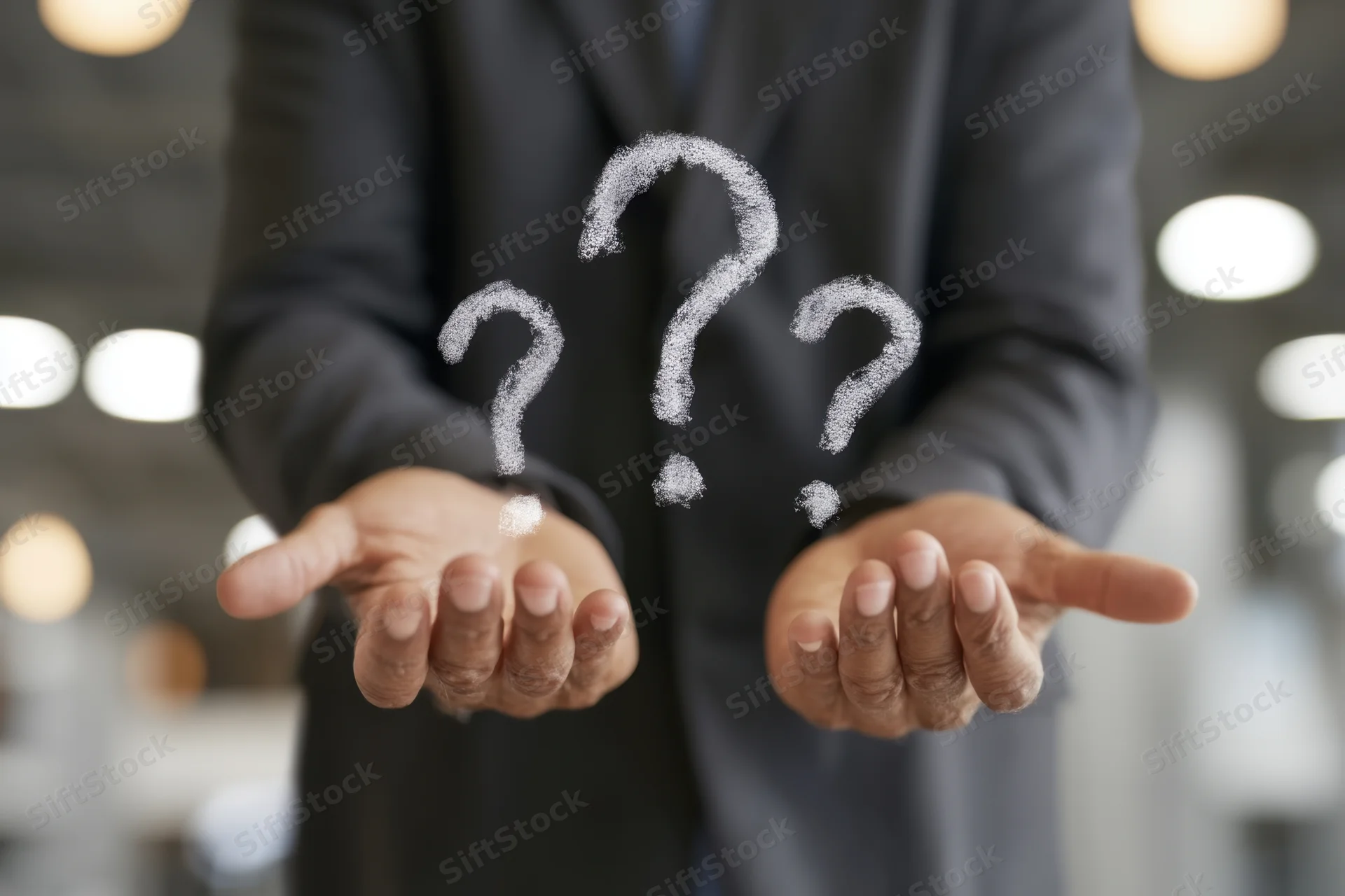 Businessman in dark suit holding open hands with floating question marks in office setting