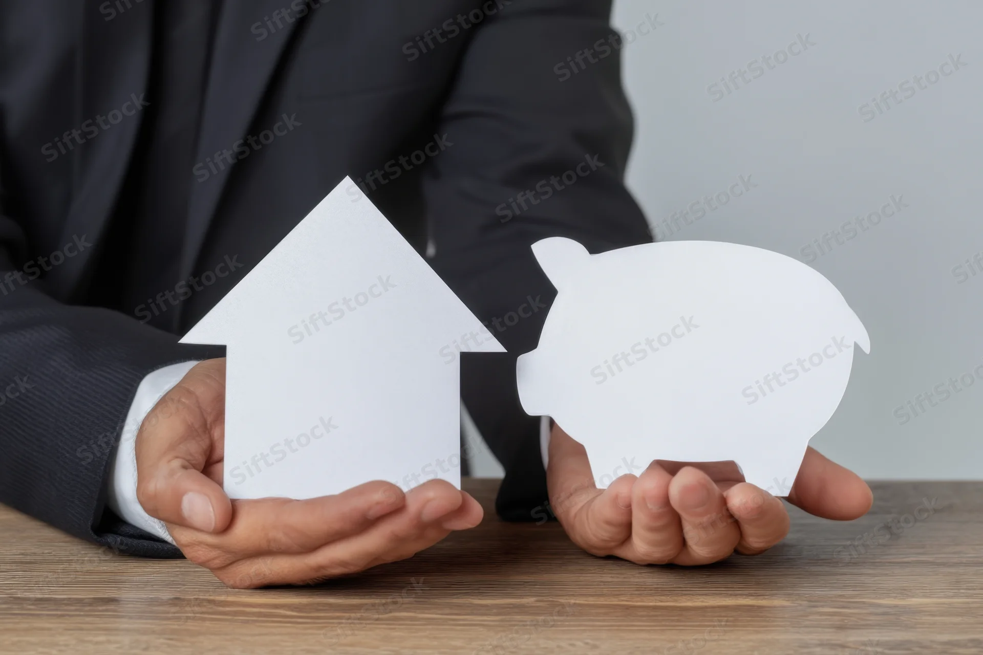 Businessman in dark suit holding white paper cutouts of a house and a piggybank on wooden table