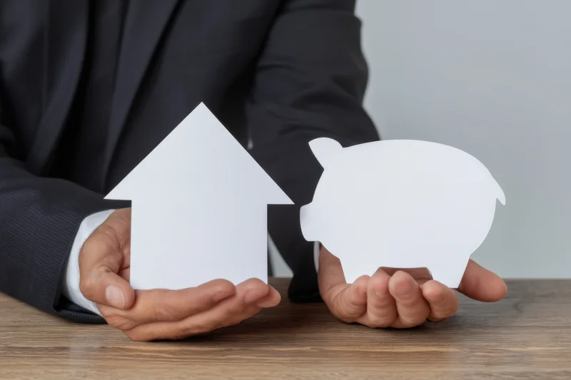 Businessman in dark suit holding white paper cutouts of a house and a piggybank on wooden table