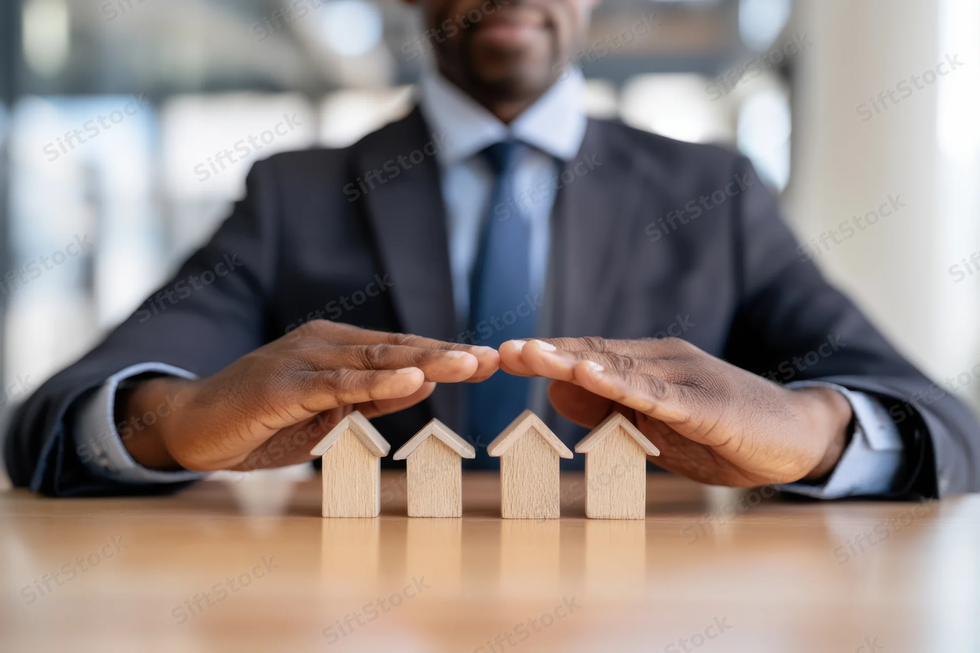 Businessman in suit placing hands over small wooden house models on desk