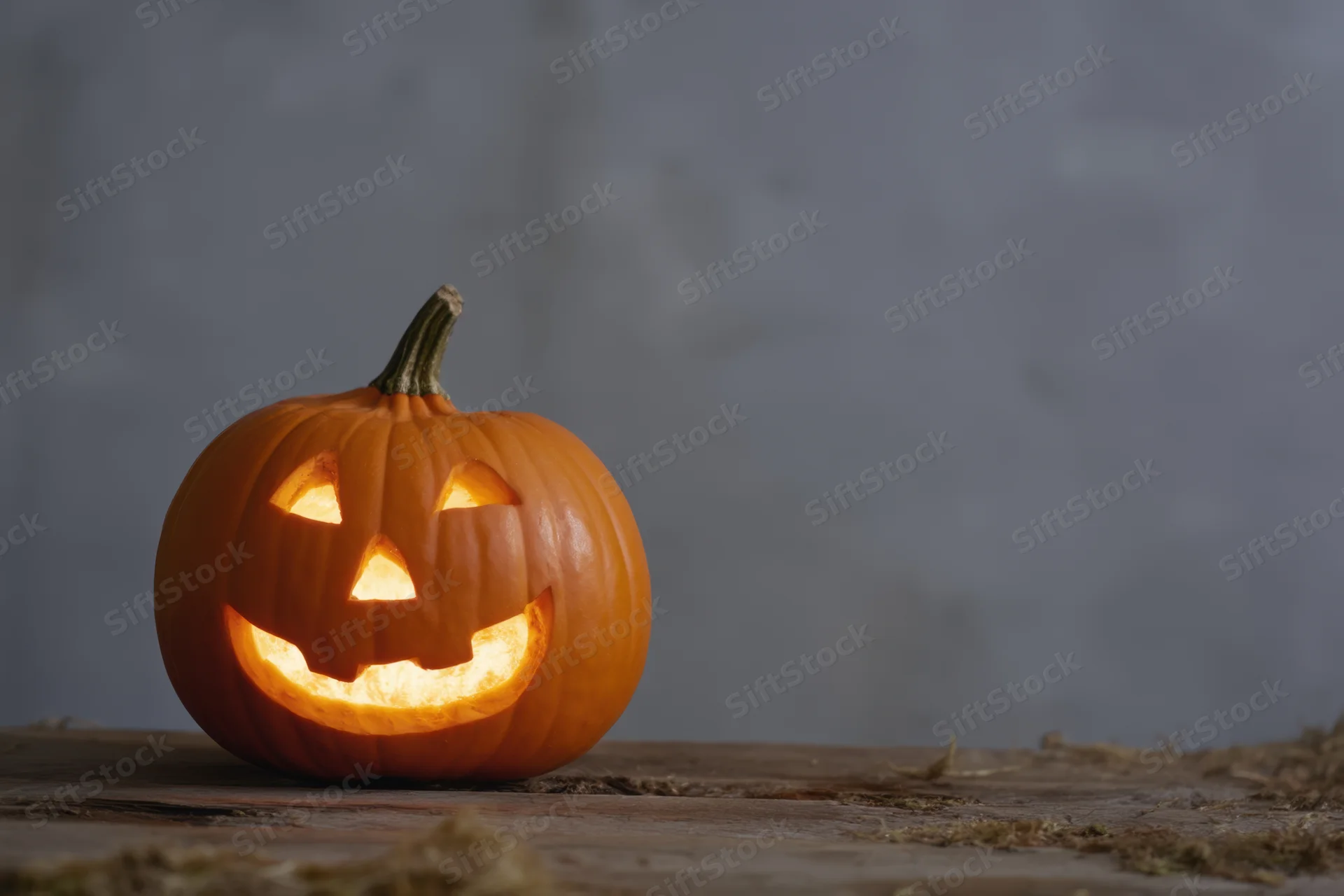 Carved pumpkin with glowing cheerful face on wooden table with hay