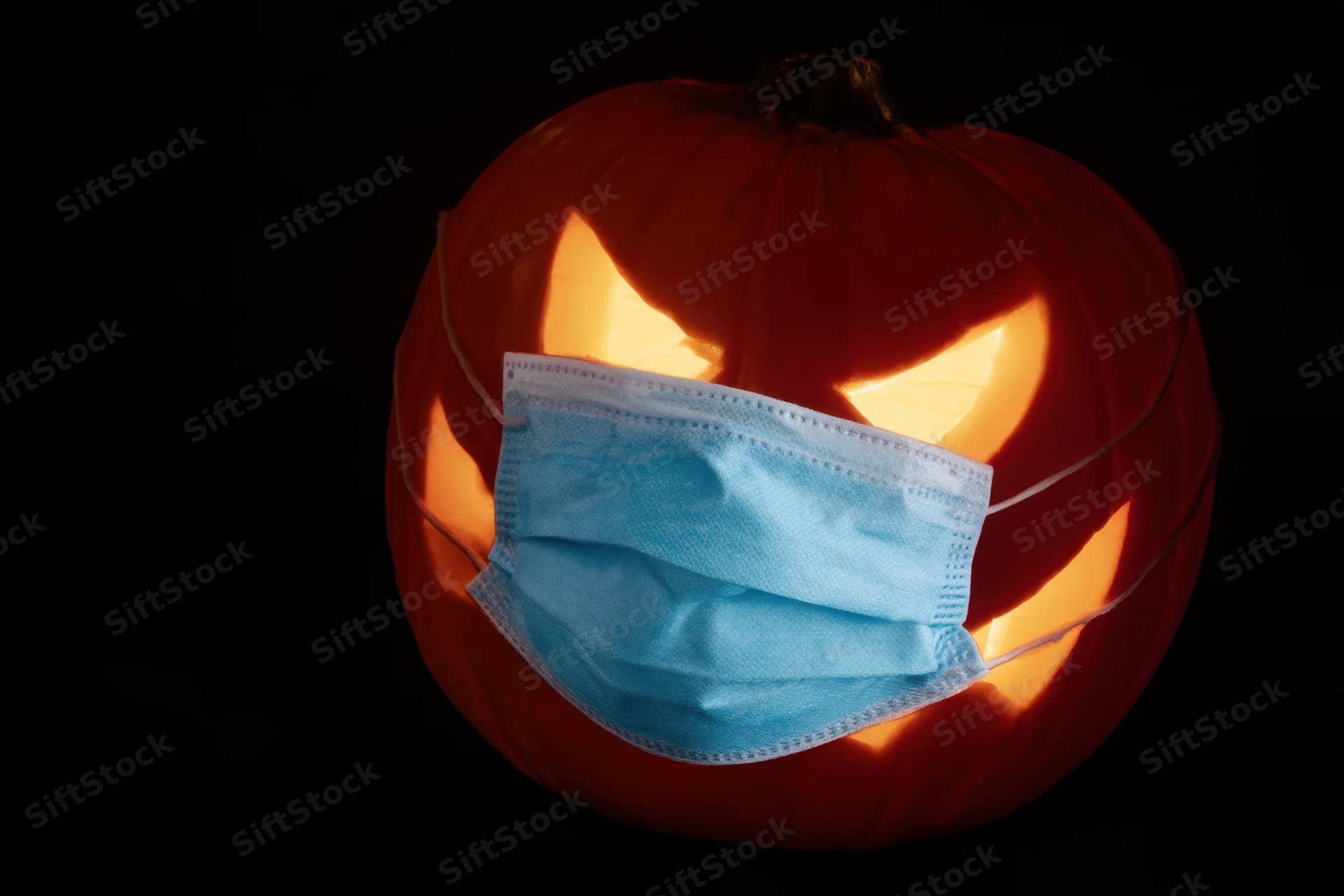 Carved pumpkin with glowing eyes and mouth wearing a blue surgical mask against dark background