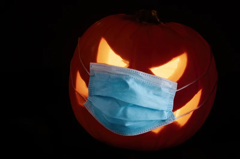 Carved pumpkin with glowing eyes and mouth wearing a blue surgical mask against dark background
