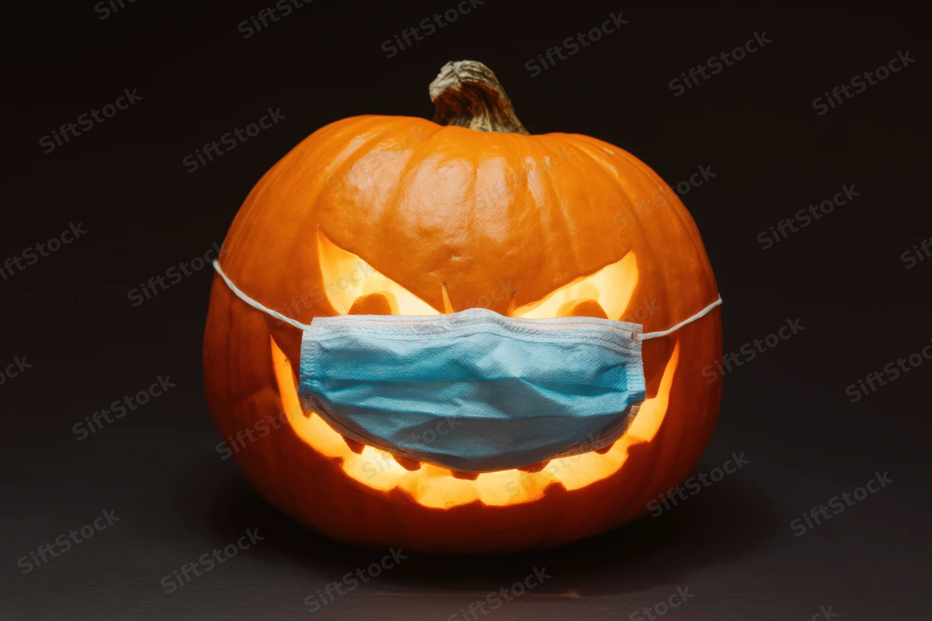 Carved pumpkin with glowing face wearing blue surgical mask against dark background