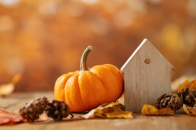 Miniature wooden house beside a small orange pumpkin on a wooden surface with autumn leaves and pinecones