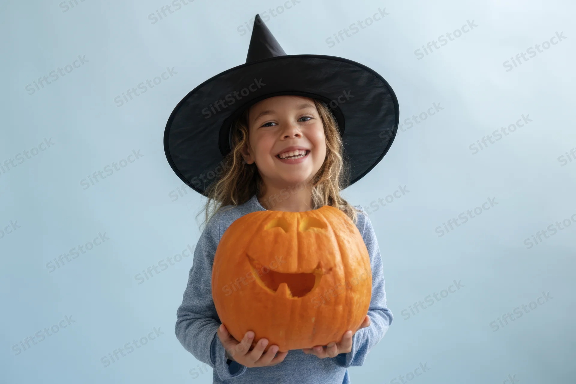 Young girl in witch hat holding carved pumpkin, smiling at camera against light blue background