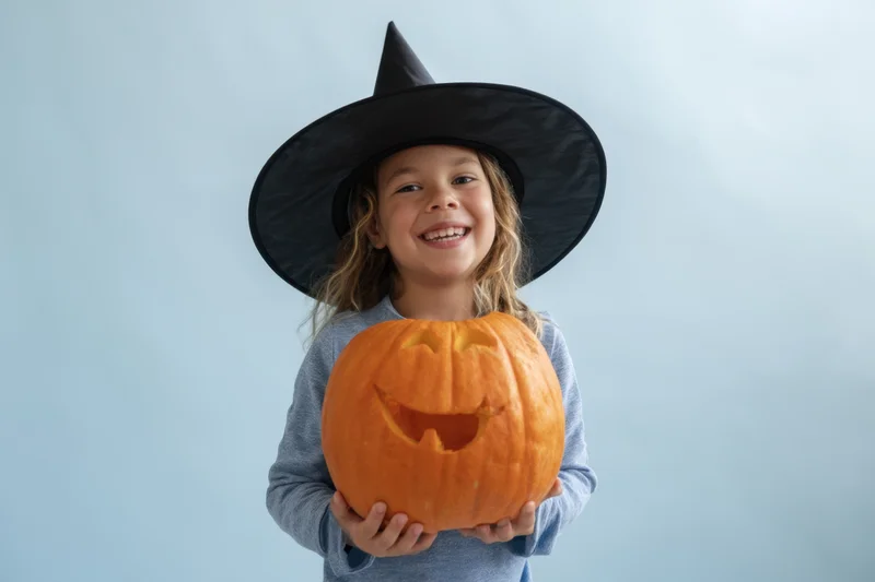 Young girl in witch hat holding carved pumpkin, smiling at camera against light blue background